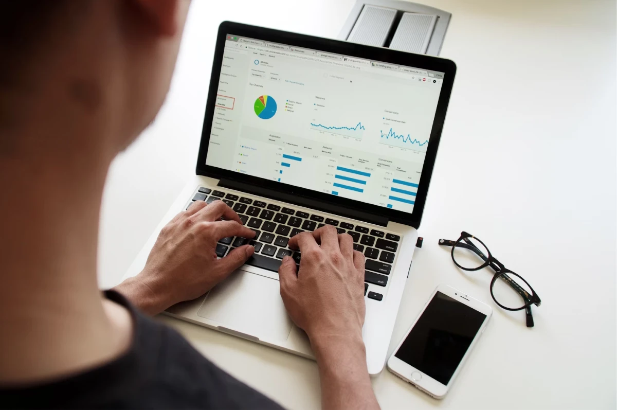 Person typing on a laptop displaying analytics charts. A pair of glasses and a smartphone lie on the desk beside the laptop.