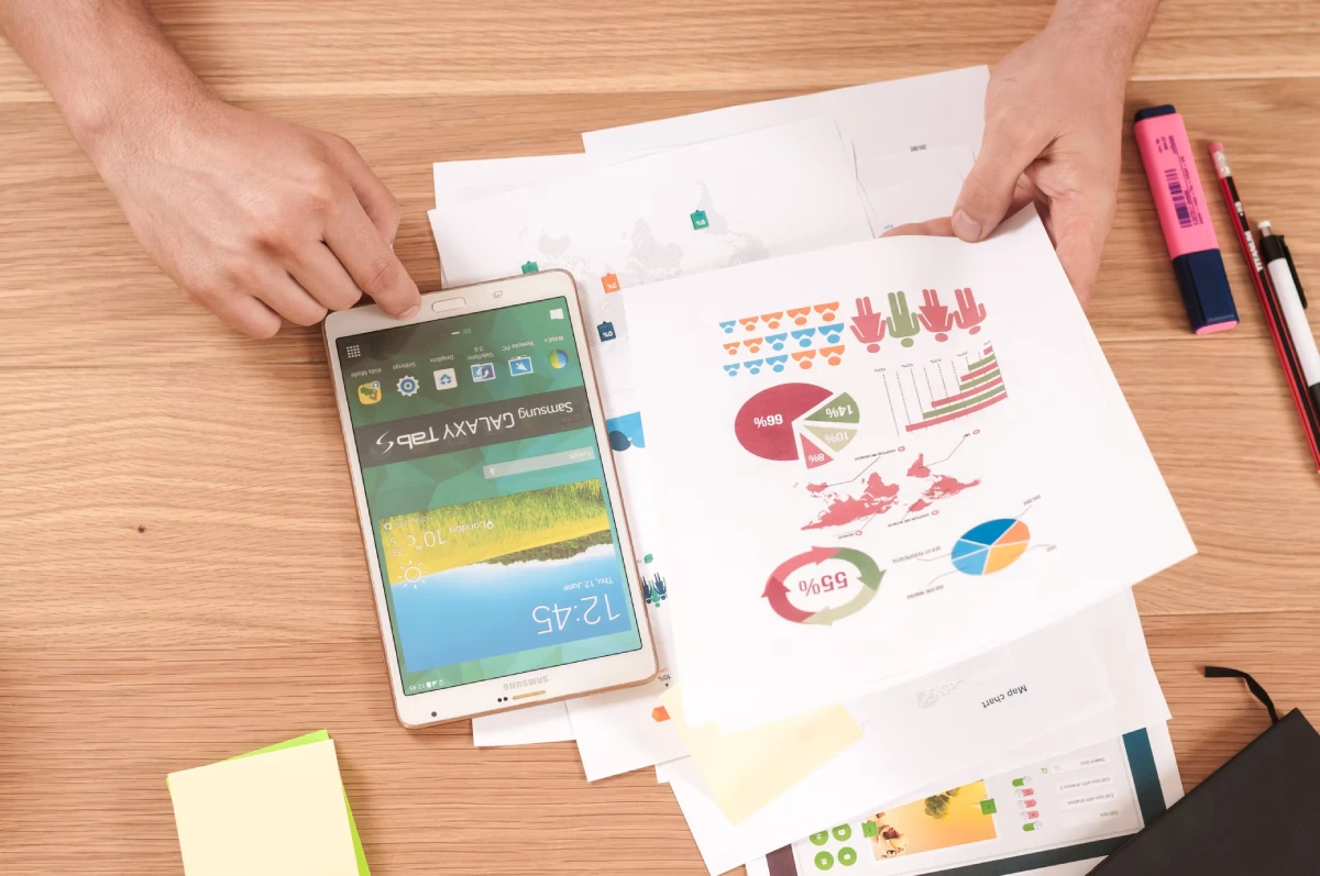 A person is analyzing charts and graphs on paper while holding a tablet on a wooden desk, surrounded by pens, a highlighter, and sticky notes.