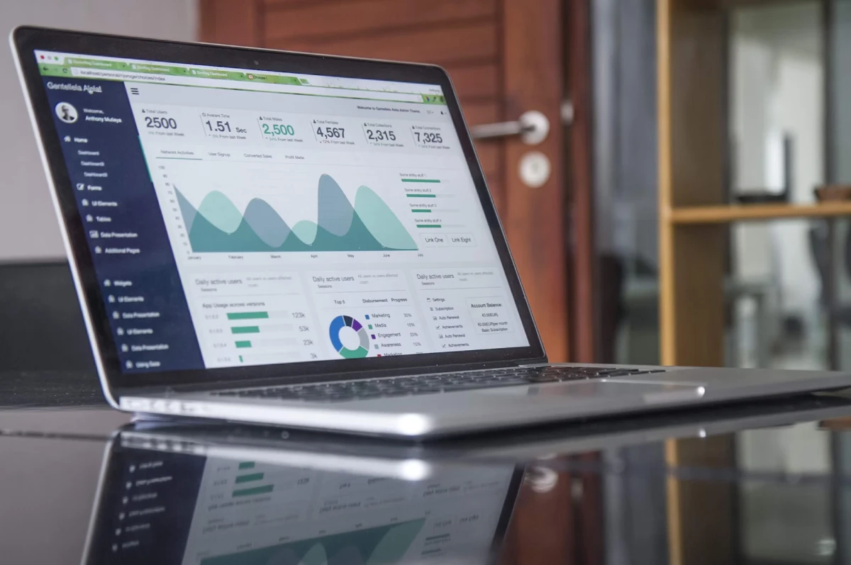A laptop displaying a data analytics dashboard with various graphs, charts, and statistics is placed on a glass table in an office setting.