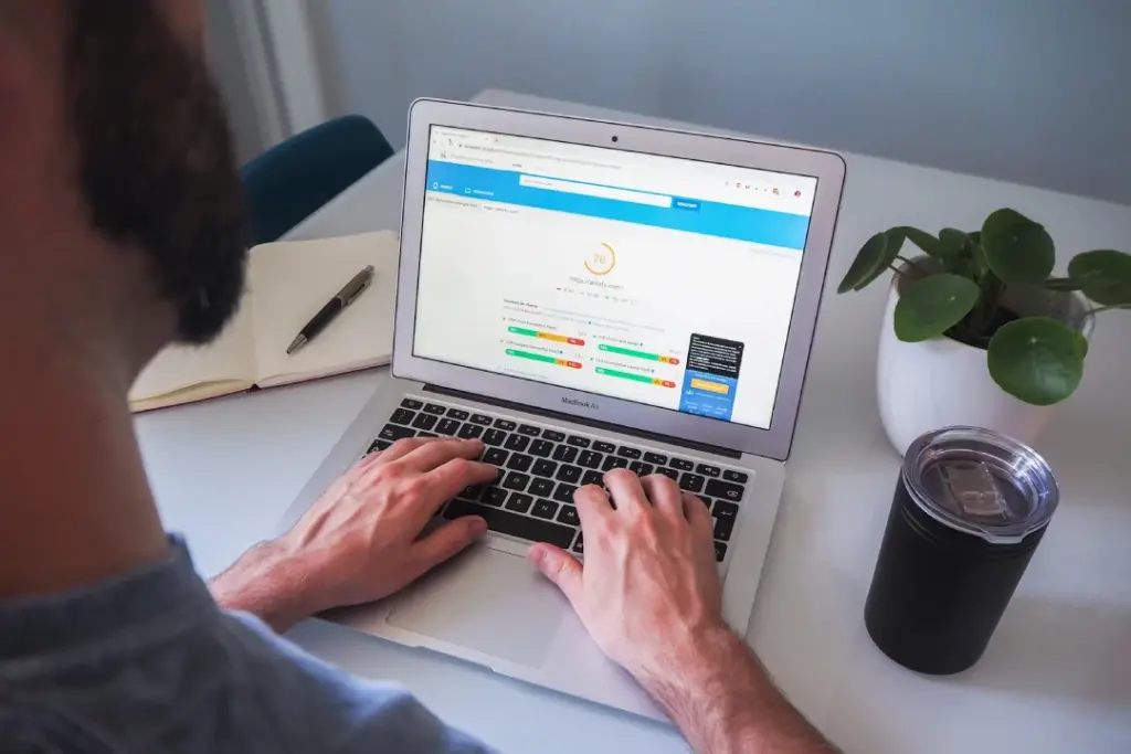 A person types on a laptop displaying an analytics dashboard. A notebook, a drink container, and a potted plant are on the desk.