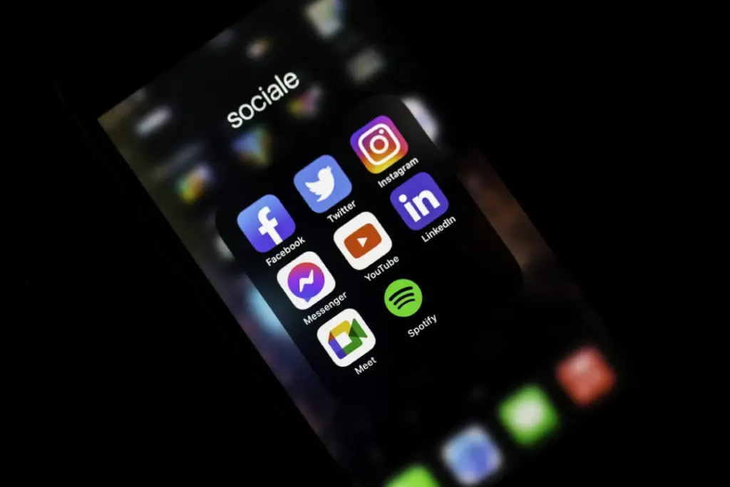 A smartphone screen displays a folder titled "sociale" containing icons for Facebook, Twitter, Instagram, Messenger, YouTube, LinkedIn, Meet, and Spotify apps against a dark blurred background.