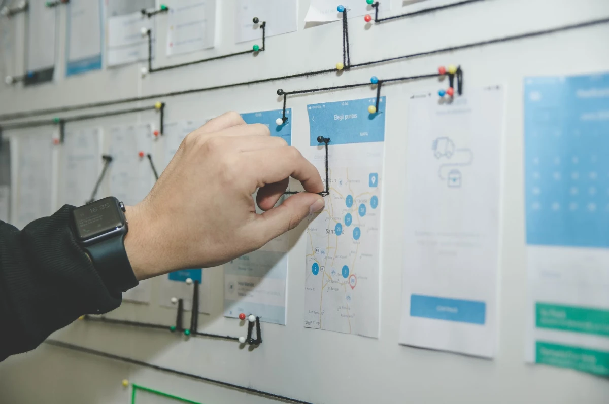 A hand wearing a smartwatch is pinning a piece of paper with diagrams on a board, surrounded by other pinned papers and strings connecting various points.